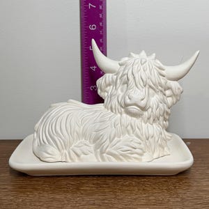 May include: A white ceramic Highland cow figurine, resting on a rectangular base. The cow has detailed fur, horns, and a focused expression. The figurine measures approximately 8 inches in length.
