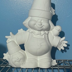 Puede incluir: Figura de cerámica blanca de un espantapájaros sonriente con un sombrero de paja, sosteniendo una calabaza y un pájaro. El espantapájaros lleva un chaleco y un mono.