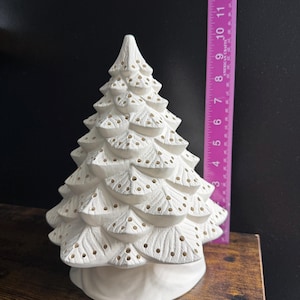 May include: A white ceramic Christmas tree figurine with a textured, tiered branch design featuring small circular cutouts. A pink ruler is positioned vertically next to the tree, indicating a height of approximately 25.4 cm from the base.