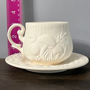 Puede incluir: Juego de taza y platillo de té de cerámica blanca con un diseño en relieve de setas y hojas. La taza tiene un asa y el platillo tiene un borde festoneado.
