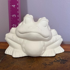 Könnte beinhalten: Weiße Keramik-Froschfigur mit glatter, abgerundeter Oberfläche. Der Frosch hat große, geschlossene Augen und ein breites, zahniges Grinsen. Er sitzt auf einer braunen Oberfläche.