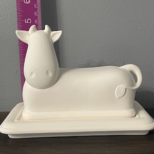 May include: White ceramic cow butter dish with a leaf design on the side. The dish is on a white ceramic base.
