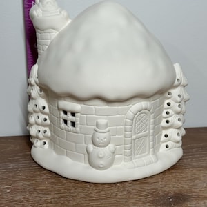 Può includere: Una casetta di Natale in ceramica bianca con tetto innevato, camino e pupazzo di neve. La casa ha una porta, una finestra e ritagli decorativi simili ad alberi. La casa è alta circa 15 cm.