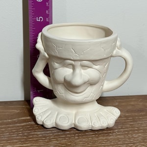 Puede incluir: Una maceta de cerámica color crema con forma de cara sonriente con cuello con volantes. La maceta tiene una nariz grande, una amplia sonrisa y un asa lateral. La maceta mide aproximadamente 13 cm de altura.