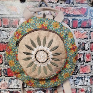 Könnte beinhalten: Ein runder, beiger Canvas-Rucksack mit einem bunten Blumenmuster und einem gestickten Sonnendesign. Der Rucksack hat zwei verstellbare Träger.