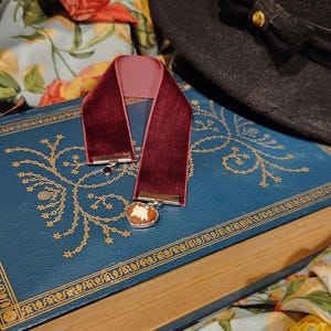 May include: A burgundy velvet ribbon with a cameo pendant rests on a blue book with gold filigree. A black hat with a bow is in the background. Floral fabric adds a touch of color to the scene.