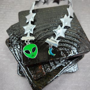 May include: Two silver-toned chokers with star accents and charms. One choker features a green alien face charm, the other a blue and green iridescent crescent moon charm. The chokers rest on a dark, textured surface.