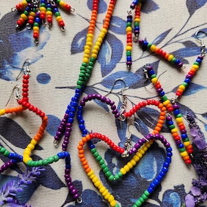 May include: A collection of handmade beaded earrings in a rainbow of colors. The earrings include various designs, such as dangling strands, heart shapes, and geometric forms. The beads are strung on silver-colored hooks.