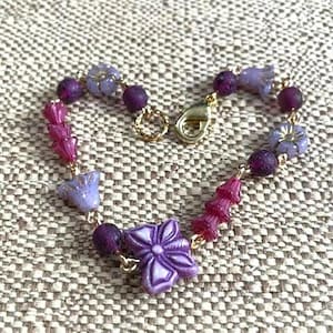 May include: A beaded bracelet featuring a central purple butterfly-shaped bead, interspersed with dark purple textured round beads, light purple flower-shaped beads, and magenta bell-shaped beads, all connected by gold-toned metal links and a lobster clasp.