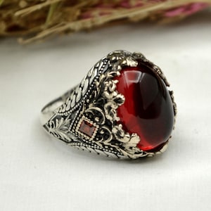 May include: A silver ring with an oval, deep red gemstone as the centerpiece. The ring features intricate, detailed patterns and a smaller square red stone on the side. The design is ornate and classic, suitable for a statement piece.