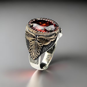 Peut inclure: Une bague en argent avec une grande pierre rouge entourée de petites pierres rouges. La bague présente des détails dorés complexes et une bande texturée.