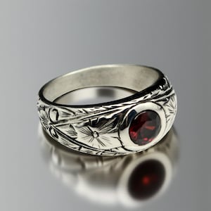 Peut inclure: Une bague en argent avec une bande gravée de motifs floraux et une pierre précieuse rouge foncé et ronde. Le design de la bague présente des motifs floraux détaillés autour de la bande.