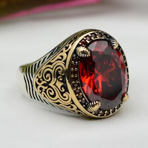 以下が含まれることがあります： 大きな楕円形の赤い宝石が付いたシルバーリング。リングは、小さな赤い石と装飾的なディテールを備えた金色のベゼルを備えています。バンドにはエッチングされたパターンと金色のアクセントがあります。