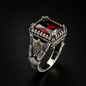 Op de afbeelding: Een zilveren ring met een grote, rechthoekige, gefacetteerde rode edelsteen. De ring heeft ingewikkelde details met gouden accenten en een decoratieve band. De ring staat tegen een donkere achtergrond.