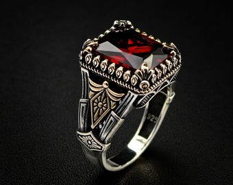 May include: A silver ring with a large, rectangular, faceted red gemstone. The ring has intricate detailing with gold accents and a decorative band. The ring is set against a dark background.