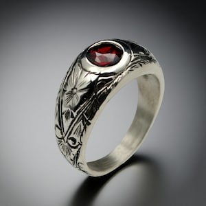 Peut inclure: Bague en argent avec une pierre précieuse rouge foncé de forme ovale. La bague présente des gravures florales et de feuilles complexes autour de l'anneau. La pierre est sertie dans un sertissage à lunette.