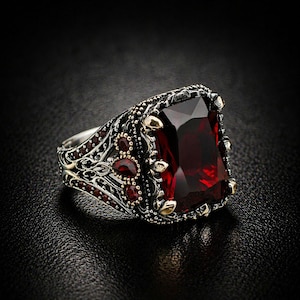 May include: A silver ring with an ornate design, featuring a large, rectangular, deep red gemstone as the centerpiece. Smaller red gemstones and gold accents adorn the ring's band, creating a luxurious and detailed appearance. The ring is set against a dark background.