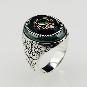 Puede incluir: Un anillo de plata con una piedra verde ovalada engastada en un bisel negro y plateado. El anillo tiene un intrincado diseño floral alrededor de la banda.