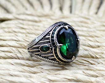 May include: A silver ring with a large, oval-shaped green gemstone surrounded by smaller green gemstones. The ring has a detailed, intricate design.