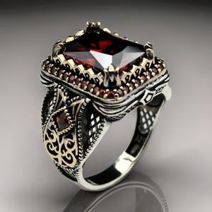 May include: A silver ring featuring a large, square, red gemstone surrounded by smaller red gemstones. The ring has intricate, ornate details and a gold band.