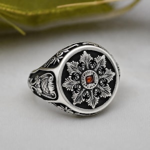 Op de afbeelding: Een zilveren zegelring met een rond voorvlak op een geoxideerde zwarte achtergrond, voorzien van een gedetailleerd, verhoogd zilveren bloem- of sneeuwvlokpatroon. Een kleine, ronde, rood-oranje edelsteen is in het midden geplaatst.