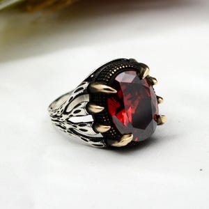 Peut inclure: Bague en argent avec une grosse pierre précieuse ovale rouge foncé. La bague a un sertissage en forme de griffes avec des accents dorés et des détails de filigrane complexes sur le bandeau.