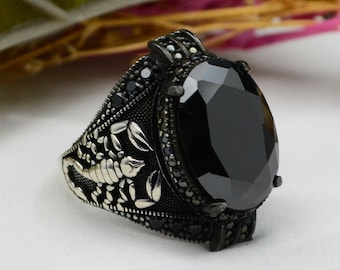 Puede incluir: Anillo plateado con una gran piedra negra de corte ovalado. El anillo presenta un diseño detallado de escorpión en la banda y piedras negras más pequeñas alrededor de la gema principal.