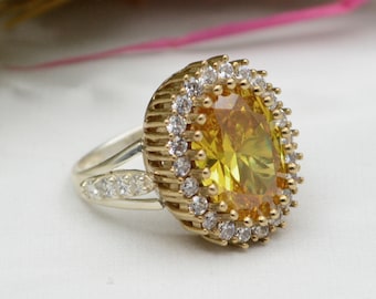 May include: A gold-toned ring featuring a large, pear-shaped yellow gemstone surrounded by a halo of smaller, clear stones. The ring's band is split, with additional clear stones set into the upper band. The ring is set against a white background.