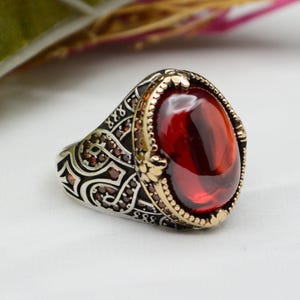 May include: A silver ring with an oval, deep red gemstone. The ring has a gold-colored bezel and intricate detailing on the band, with small red accent stones. The design is ornate and eye-catching, suitable for a statement piece.