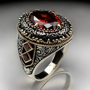 May include: A silver ring with intricate floral designs and a large red gemstone. The ring is accented with gold details and smaller red gemstones.