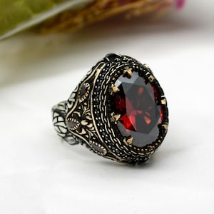 Puede incluir: Un anillo de plata adornado con una gran piedra preciosa roja oscura facetada de forma ovalada. El anillo presenta intrincados filigranas y detalles negros alrededor de la piedra preciosa y en la banda. También hay pequeños detalles dorados.