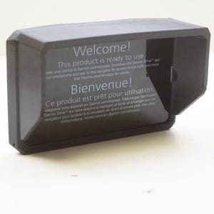 Puede incluir: Un dispositivo rectangular negro con lados angulados, con el texto "Welcome! This product is ready to use." y "Bienvenue!" en blanco. El dispositivo parece ser un soporte o una cubierta para un dispositivo electrónico.