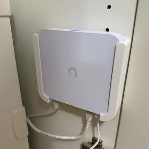 May include: A white rectangular electronic device with a curved logo is mounted on a white bracket. Two white cables are connected to the device. The device is mounted on a white surface.