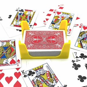 Pode incluir: Um porta-cartões de plástico amarelo com um baralho de cartas dentro. O porta-cartões está rodeado de cartas de jogo com diferentes naipes e números.