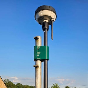 Puede incluir: Un dispositivo de topografía para exteriores con una parte superior en blanco y negro, un poste negro y un soporte de montaje verde. El dispositivo tiene una pequeña antena y se encuentra contra un cielo azul claro.
