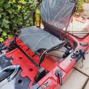 May include: A red kayak with a black and gray seat. The seat is made of a mesh material and has adjustable straps. The kayak also has a black frame and various accessories.