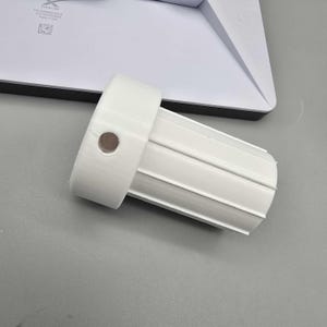 May include: A white plastic cylindrical component with a ridged body and a circular hole. The component is positioned on a gray surface, with a white rectangular object in the background.