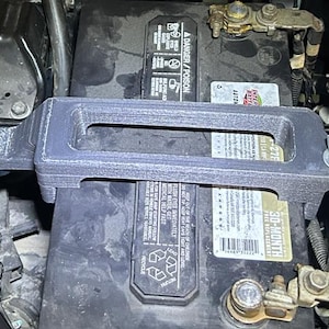 May include: A dark gray battery hold-down clamp installed over a black car battery. The clamp has a rectangular shape with a handle and is secured with a bolt. The battery has warning labels and a 30-month free replacement sticker.