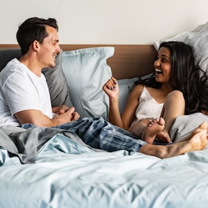Puede incluir: Una pareja en la cama riendo y hablando entre ellos. Ambos llevan pijamas y están rodeados de almohadas. La cama tiene una funda nórdica azul claro y sábanas blancas.