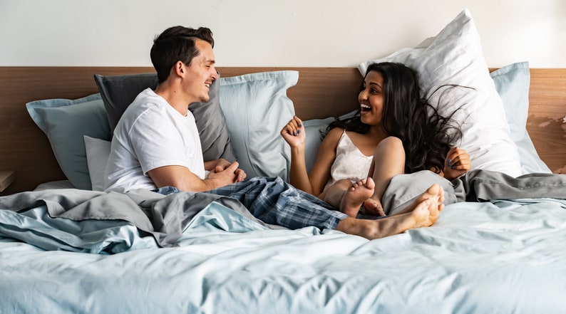 K&ouml;nnte beinhalten: Ein Paar im Bett, das lacht und miteinander spricht. Beide tragen Schlafanz&uuml;ge und sind von Kissen umgeben. Das Bett hat einen hellblauen Bettbezug und wei&szlig;e Laken.