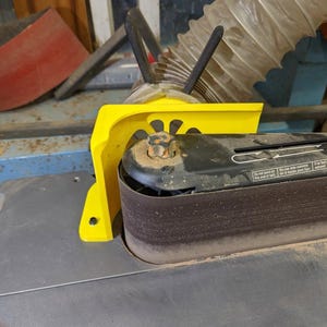 May include: A yellow safety guard attached to a black and gray belt sander. The sander has a brown sanding belt and a metal base. The guard is made of bright yellow plastic and is designed to protect the user from the moving belt.