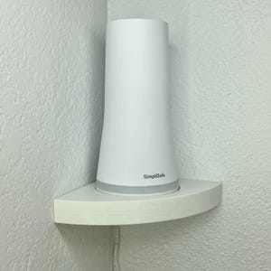 May include: A white SimpliSafe security system base station on a white corner shelf. The base station is cylindrical with a flared base and a silver accent. The shelf is mounted on a textured white wall.