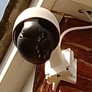 Peut inclure: Une caméra de sécurité blanche avec un objectif noir montée sur un support blanc. La caméra est montée sur un mur en briques.