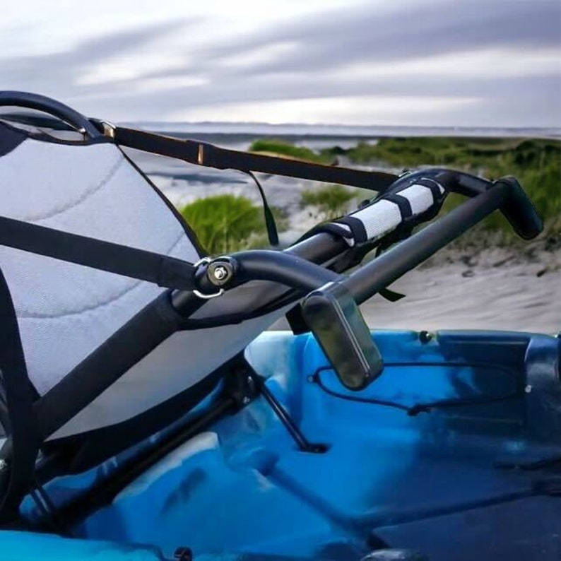May include: A close-up of a kayak seat with a black frame and a light gray and white seat. The kayak is blue. The seat has black straps and a black handle. The background is a beach with the ocean in the distance.