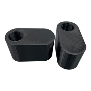 May include: Two black, 3D-printed, rectangular objects with rounded edges. Each object has a cylindrical hole on top. The objects are made of a textured material and are set against a white background.