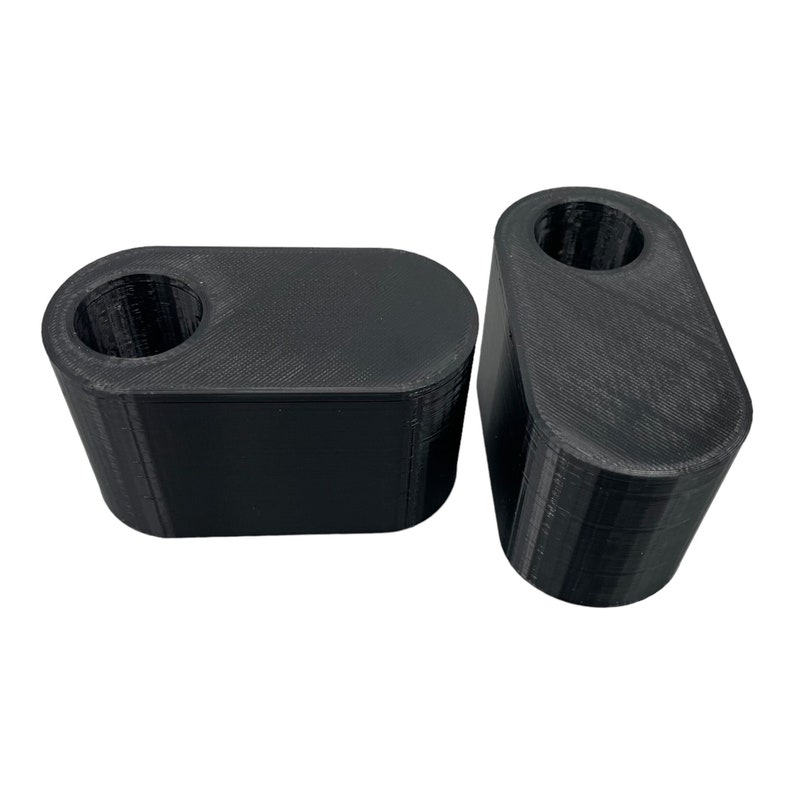 May include: Two black, 3D-printed, rectangular objects with rounded edges. Each object has a cylindrical hole on top. The objects are made of a textured material and are set against a white background.