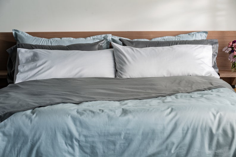 K&ouml;nnte beinhalten: Ein Bett mit einem hellblauen Bettbezug und einem grauen Bettbezug, der am Fu&szlig;ende des Bettes zusammengefaltet ist. Zwei wei&szlig;e Kissen und zwei graue Kissen liegen auf dem Bett.