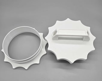 May include: Two-piece white plastic cookie cutter set. One piece is a circular ring with a scalloped edge, and the other is a flat, sunburst-shaped piece with a handle. The set is designed for baking.