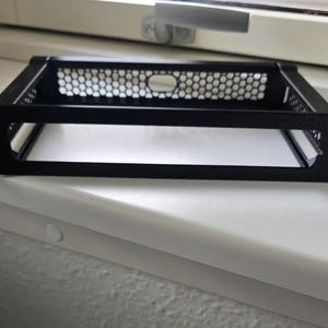 Puede incluir: Un componente de montaje en rack de metal negro con un diseño de malla de panal. El marco rectangular tiene orificios de montaje en la parte delantera y está diseñado para contener equipos electrónicos. El componente está sobre una superficie blanca.