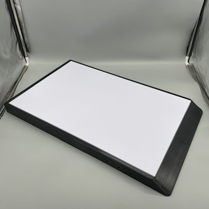May include: A black rectangular light box with a white surface. The light box has a beveled edge and is designed to be used for displaying artwork or other objects.